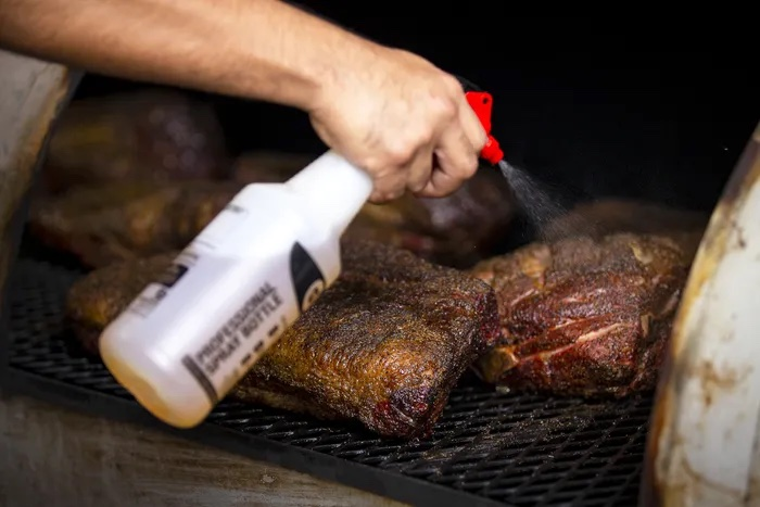 A person managing smoke and moisture in a smoker grill by spraying meat with apple juice